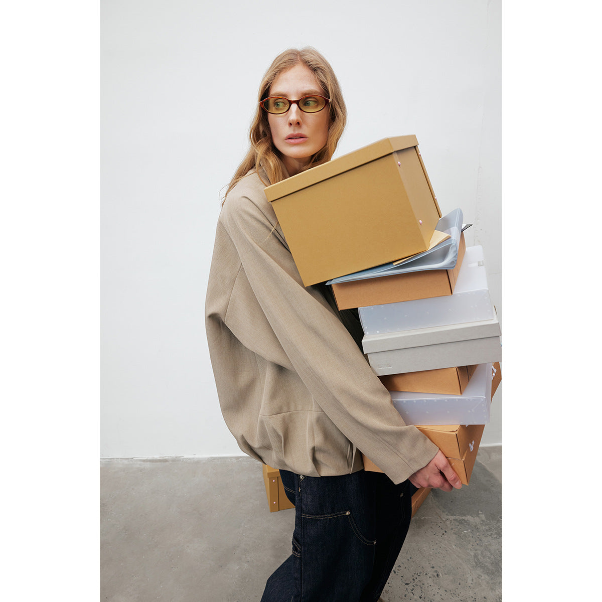 Model in a taupe jacket and dark jeans carries a stack of cardboard and white boxes.