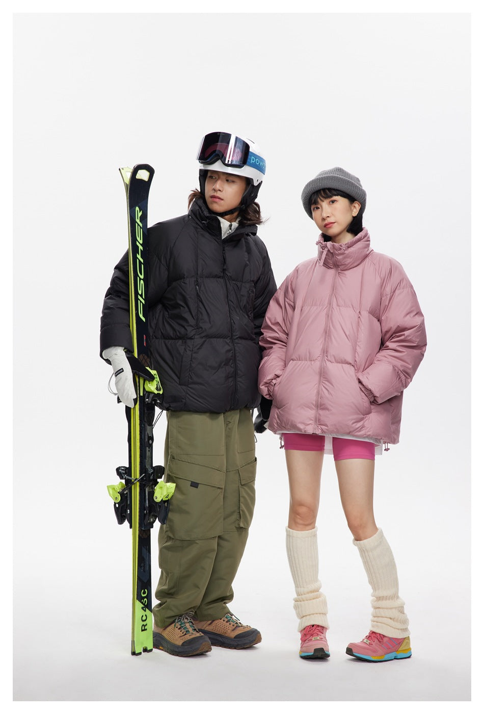 Two models wear black and dusty pink puffer jackets with ski gear against a white studio background.