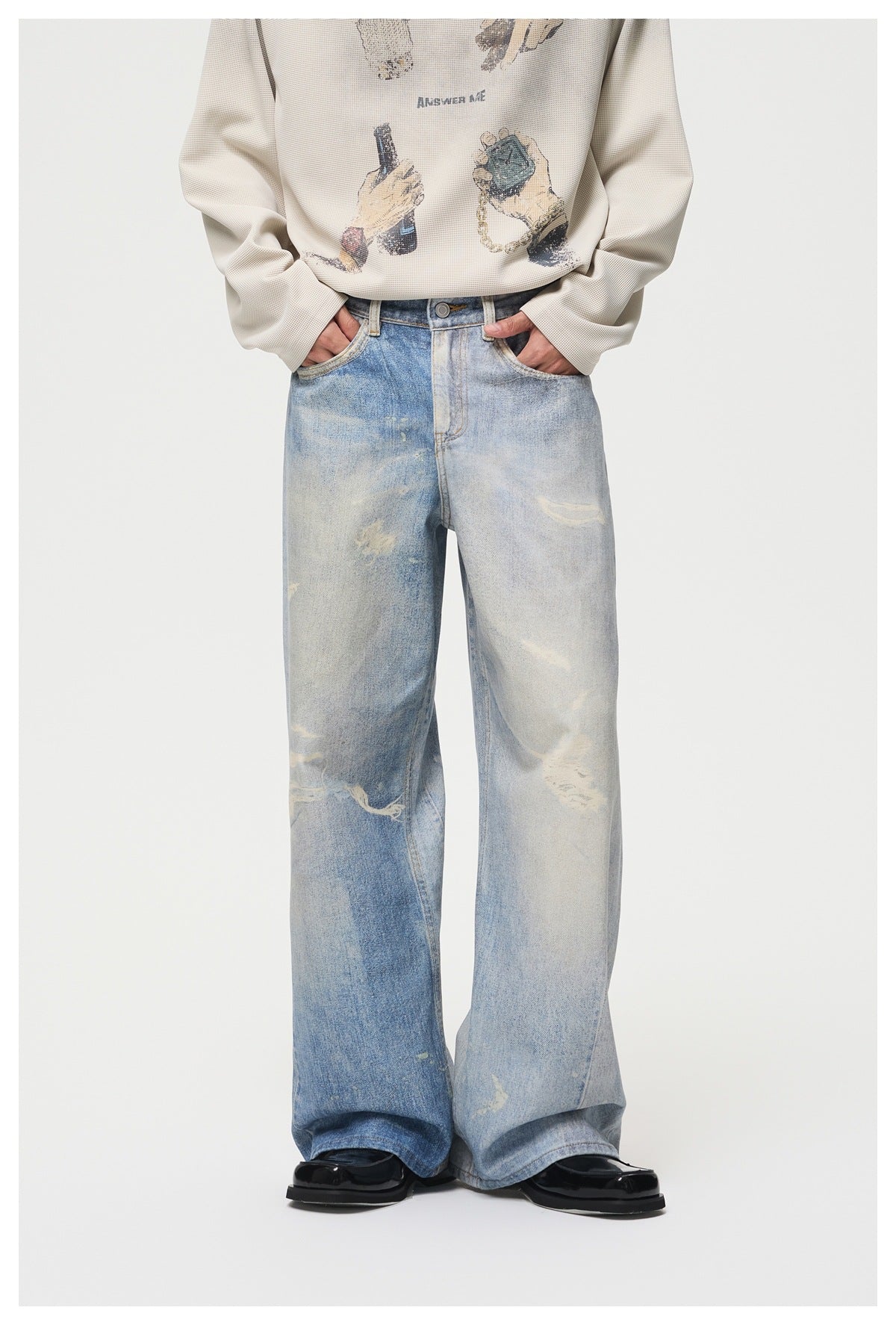 Model wearing light blue distressed wide-leg jeans with hands in pockets, a beige printed sweatshirt and black shoes