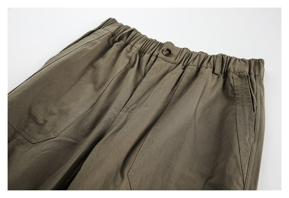 Close-up of olive trousers showing the gathered waistband, button closure and front pocket construction.