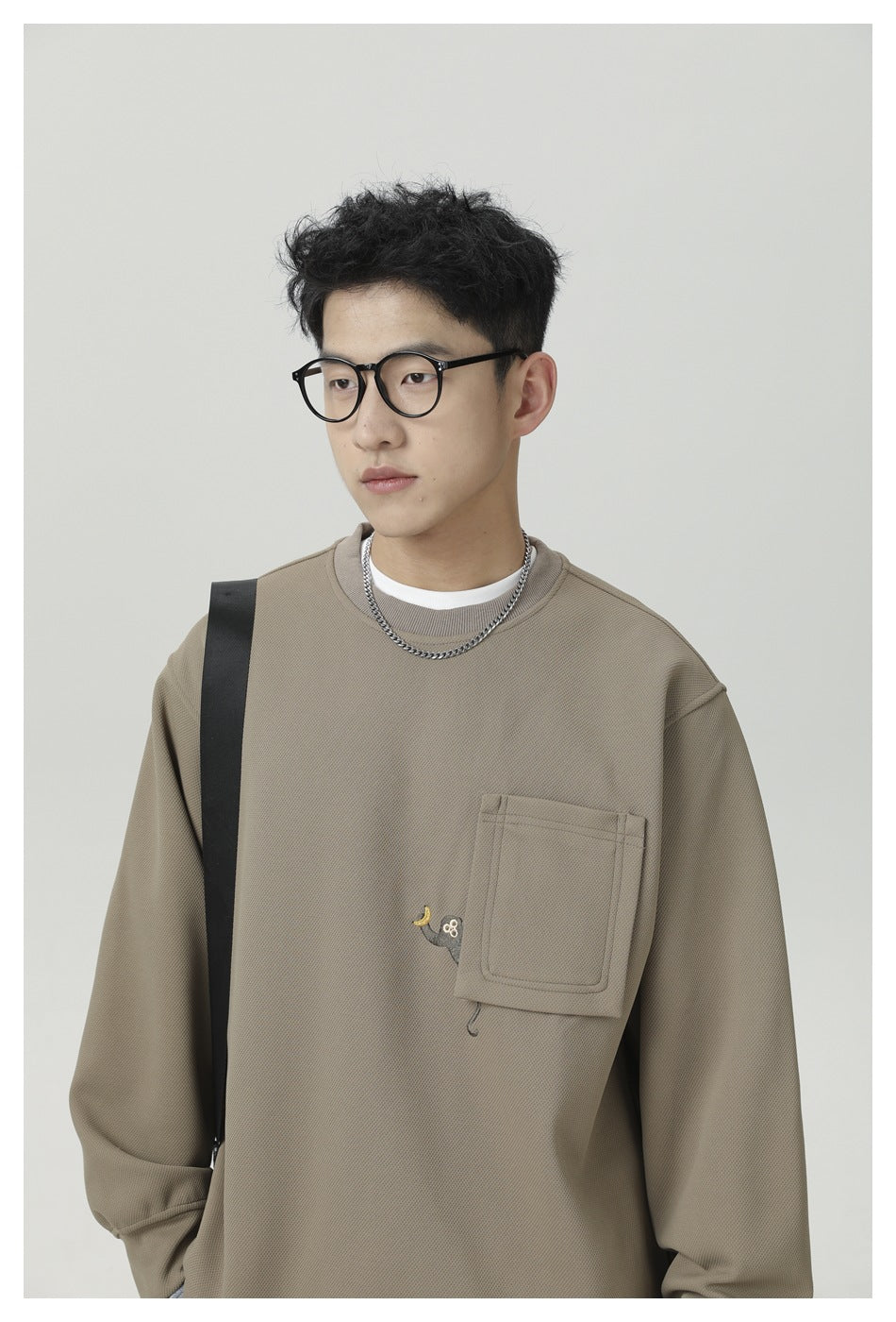 Upper-body view of a model wearing a taupe crewneck sweatshirt with a large chest pocket and small embroidered motif.