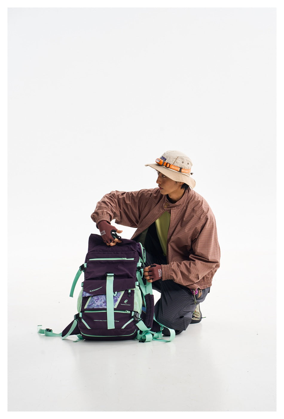 Model crouching beside a large purple and mint backpack, wearing a rose checked jacket and dark trousers.