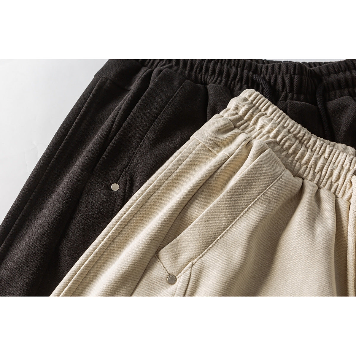 Close-up of black and beige pants showing elastic waistbands, pleats, angled pockets and small metal rivets.