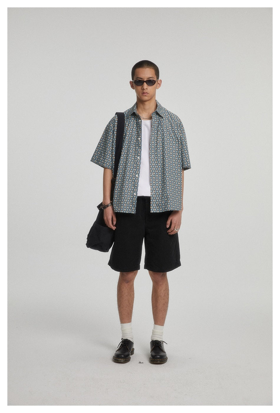 Model wearing a blue patterned short-sleeve shirt over a white top with black shorts.