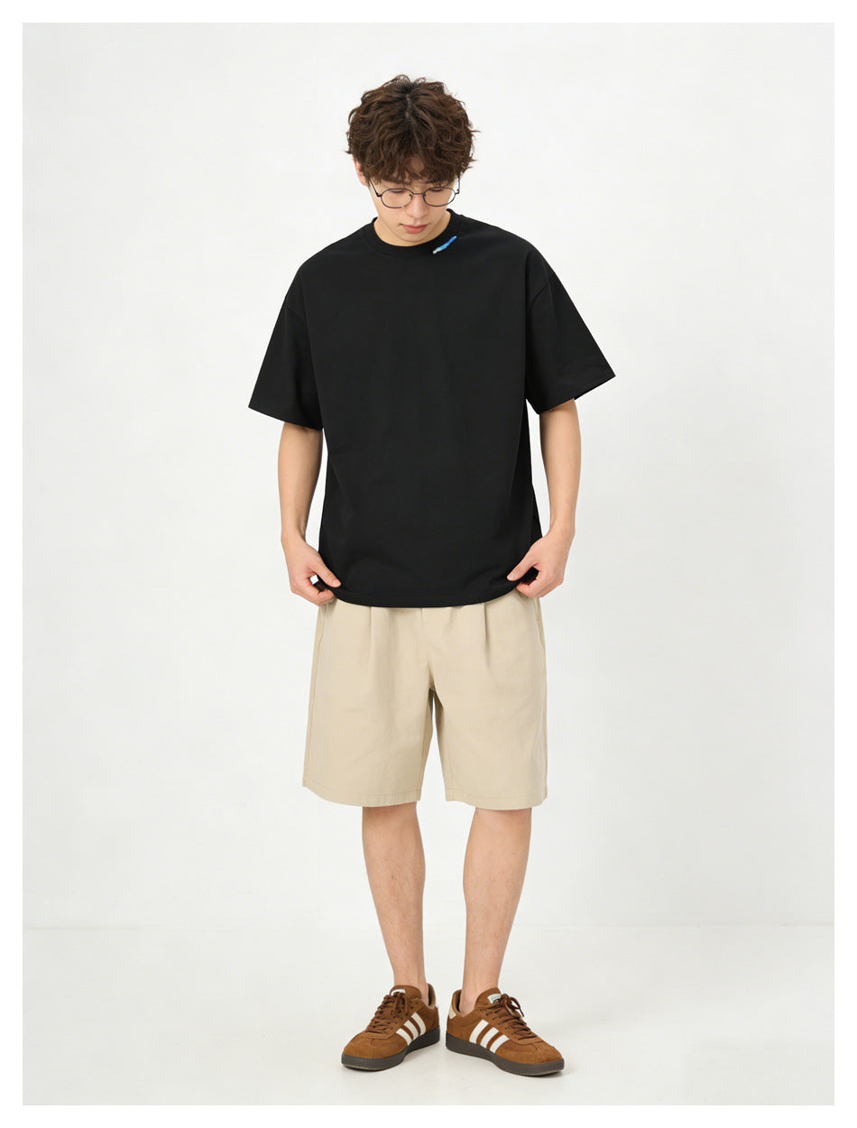 Man wearing black relaxed t-shirt with blue collar embroidery, beige shorts and brown sneakers, looking down