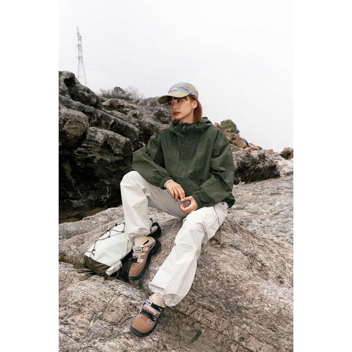 A seated woman outdoors wears a dark olive hooded jacket, light trousers and a gray cap.