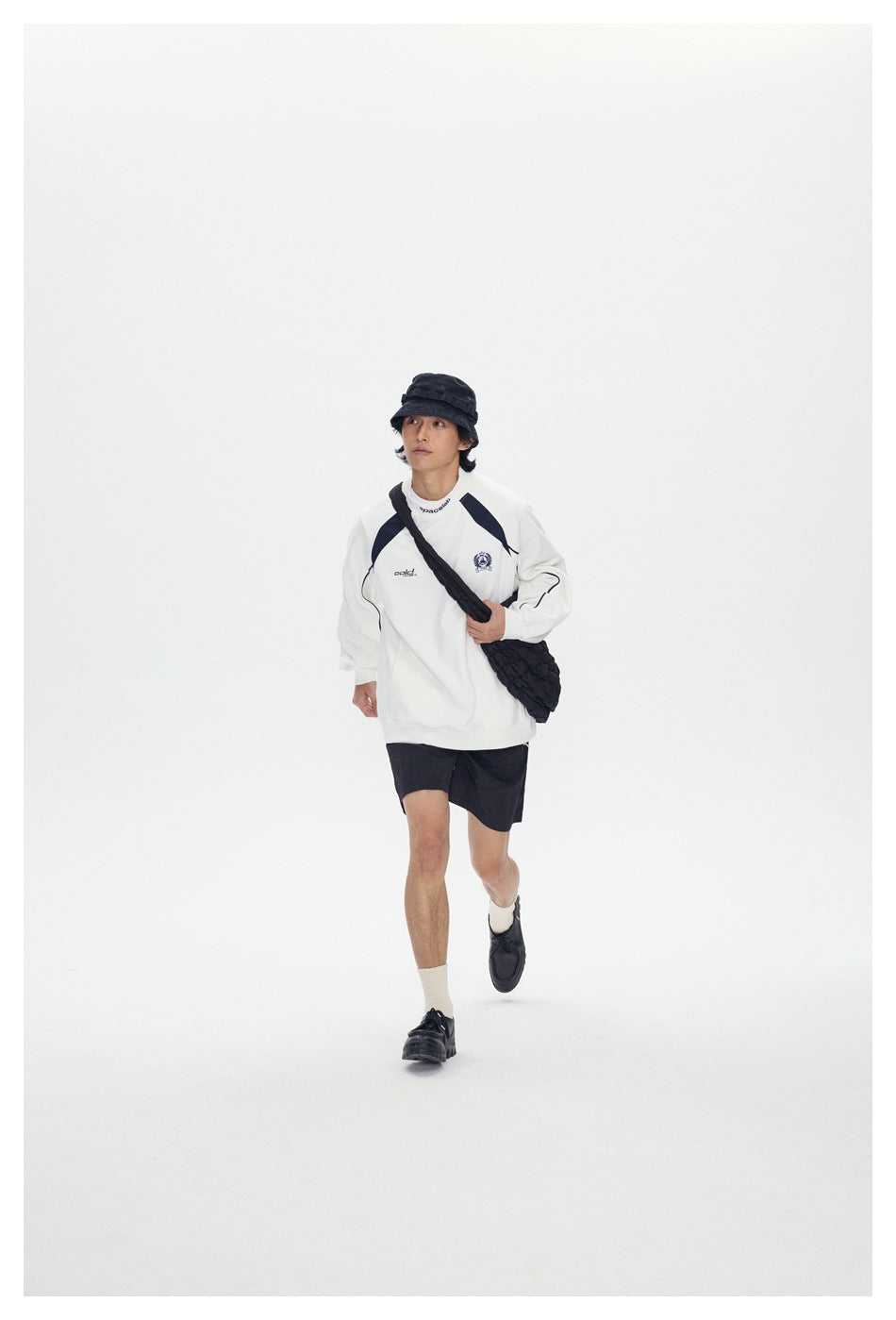Model walking in a white graphic sweatshirt and black shorts with a black bucket hat and crossbody bag.