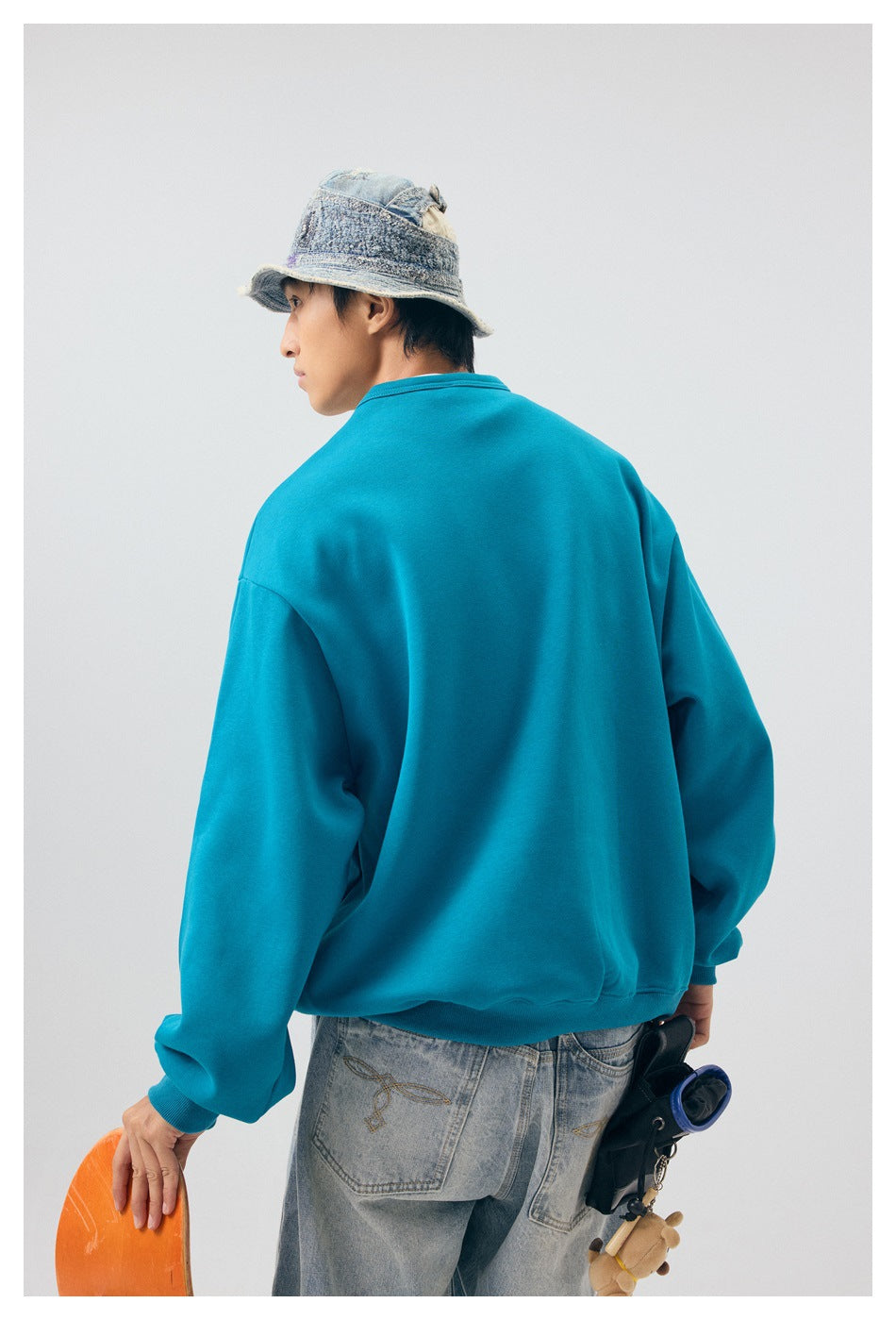 Model wearing the plain back of a teal sweatshirt, shown from behind with jeans and a bucket hat.