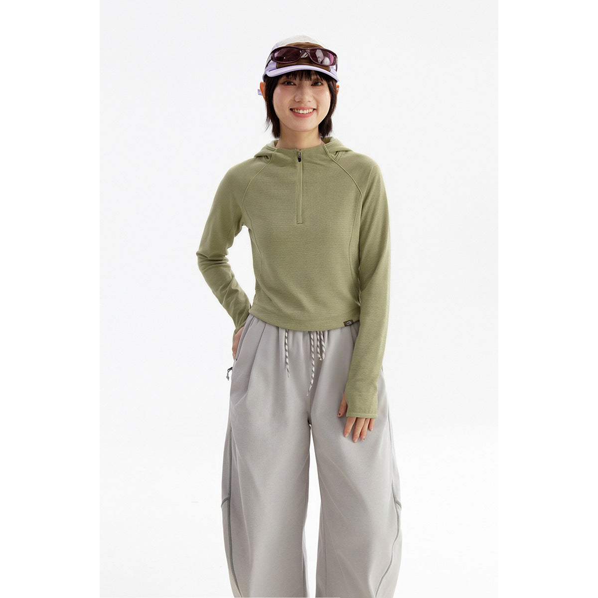 Model wearing a sage green hooded pullover with a short zip neck and loose light gray pants.