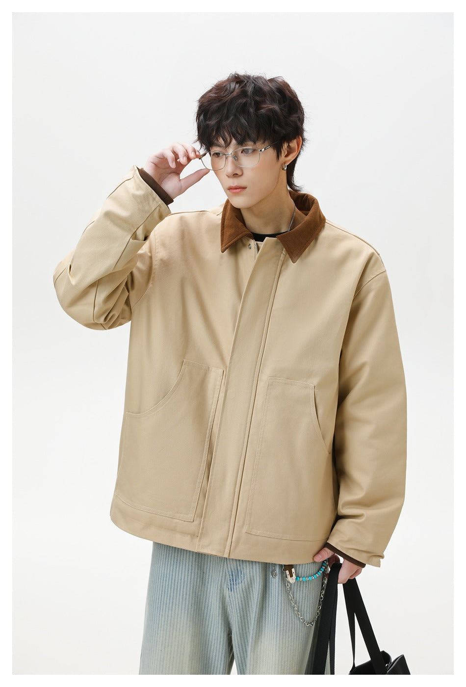 Model adjusts his glasses while wearing a beige jacket with a brown collar and large front pockets.