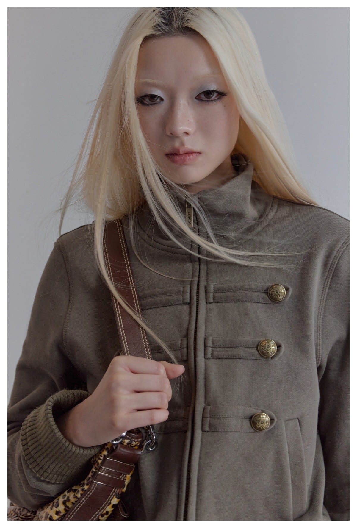 Close-up of a model wearing the grey-green jacket, showing the stand collar, gold-tone buttons, pocket and ribbed cuff