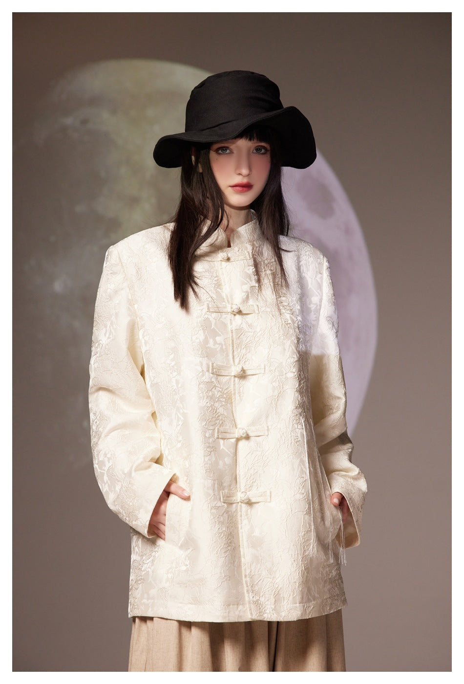 Front view of a model wearing an ivory floral-textured jacket with frog closures and a black wide-brim hat.