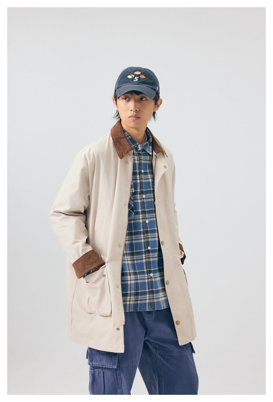 Model wearing an open light beige coat with brown collar and cuffs over a blue plaid shirt and navy cargo pants.