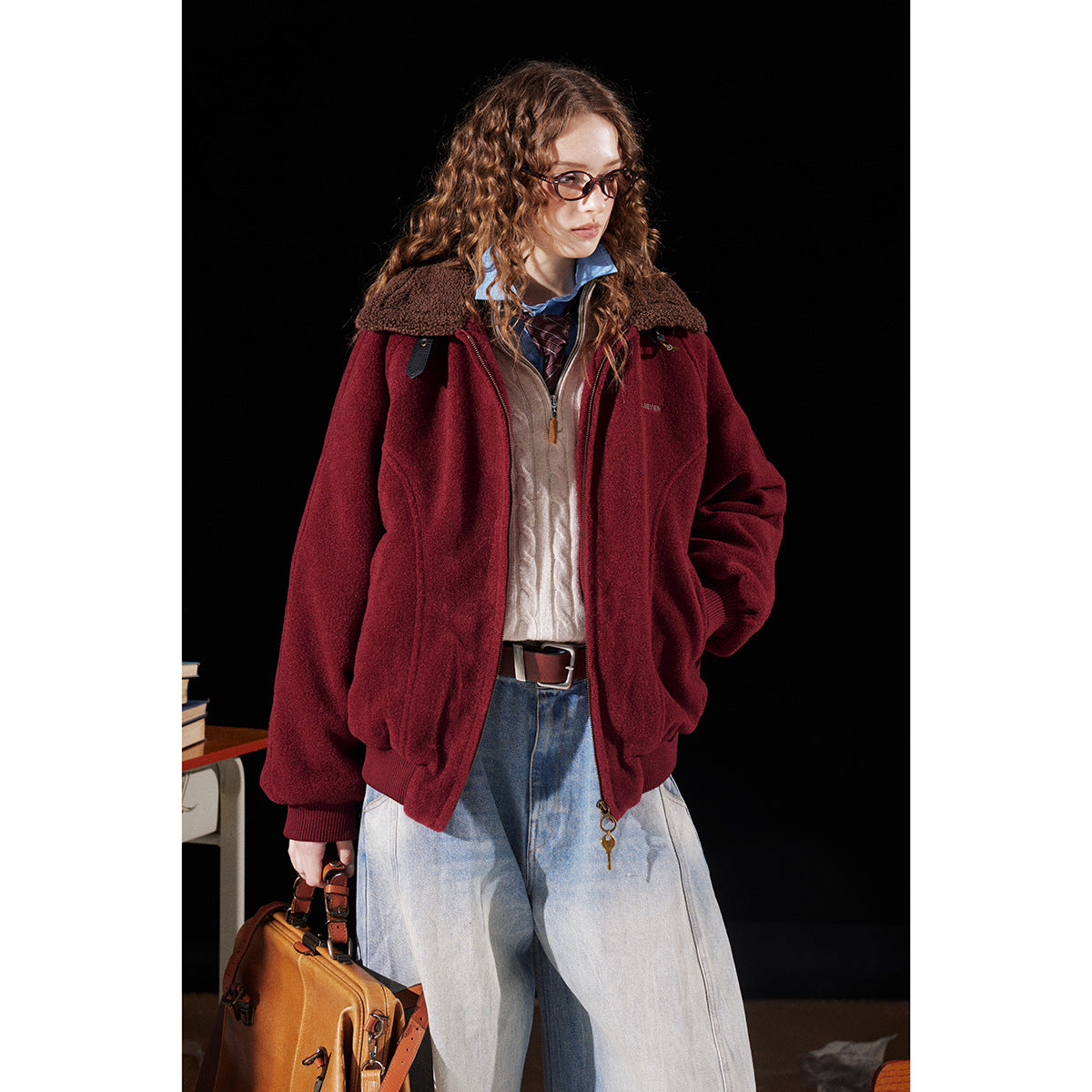 Model wearing a burgundy zip jacket with a brown sherpa collar, layered over a cream sweater and light jeans.