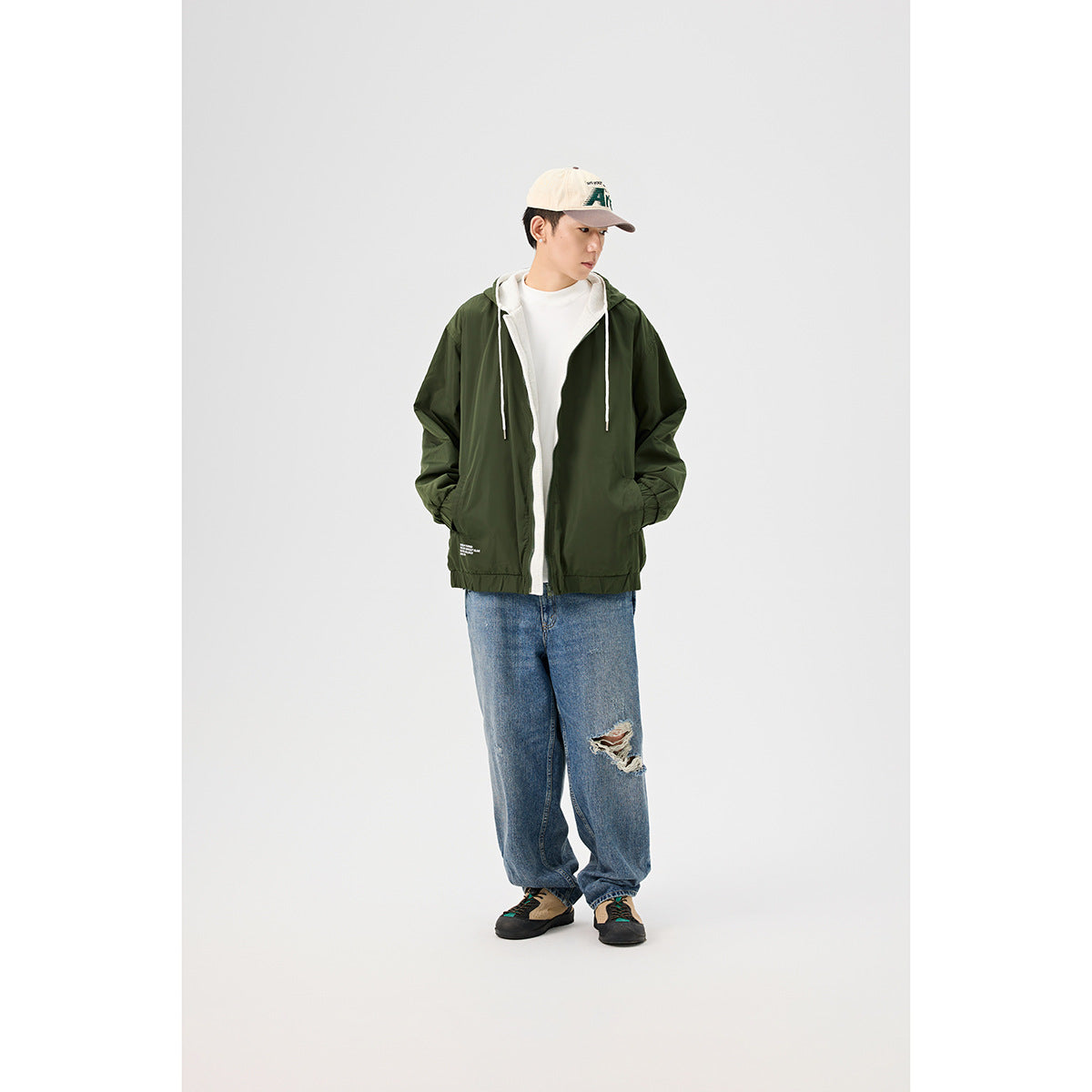 Model wearing a dark olive hooded zip jacket over a white shirt, with loose blue jeans and a cap.