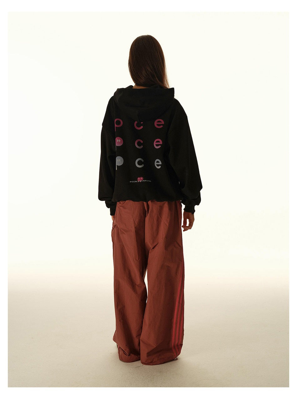 Back view of a model wearing a black hoodie with multicolored lettering and a small graphic, with rust wide-leg pants.