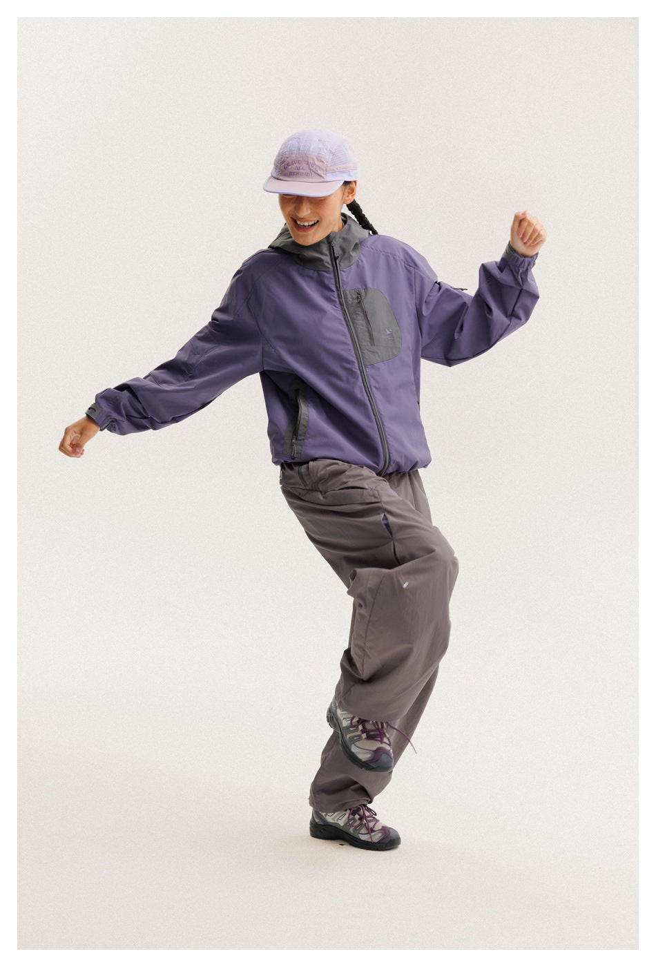 Model in a purple hooded jacket with charcoal panels and loose gray pants, posing in a lavender cap.