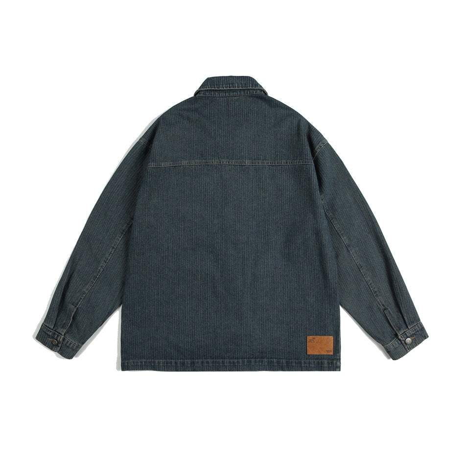 Back view of a dark blue denim jacket laid flat, with a pointed collar, long sleeves, and a small brown hem patch.