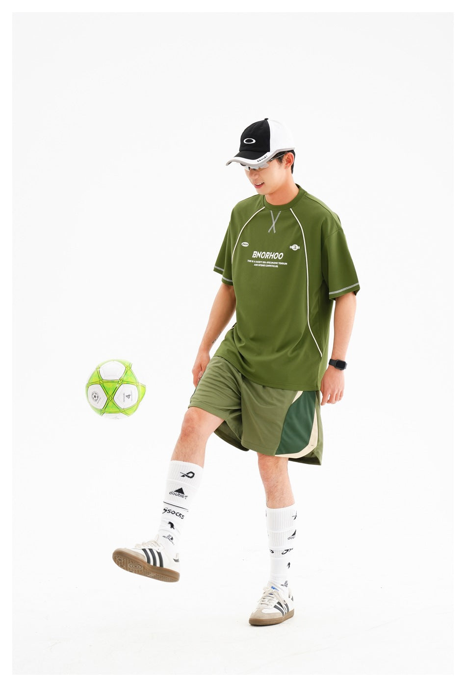 Man in an olive-green sports T-shirt and matching shorts, kicking a green-and-white soccer ball.