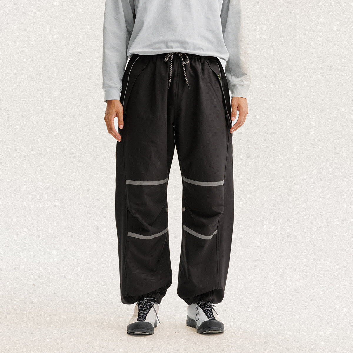 Front view of a model wearing loose black drawstring pants with gray reflective bands and gathered ankles.