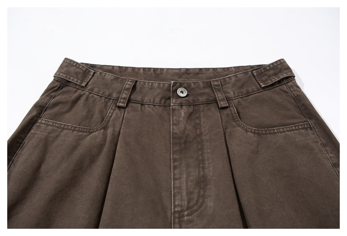Close-up of brown trousers showing the waistband, button, belt loops, front pleats and curved pockets.