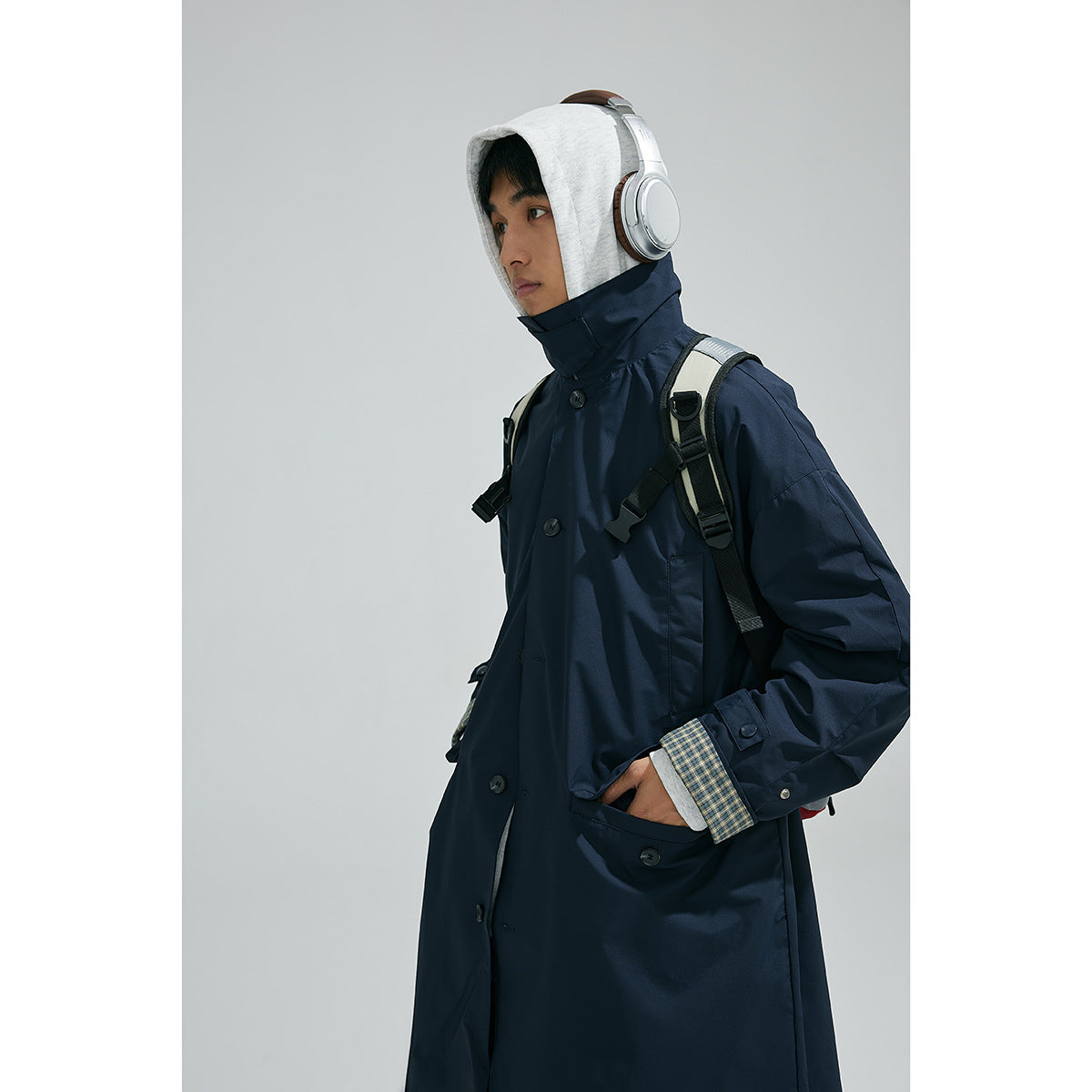 Model wearing a long navy coat with a high collar, plaid cuff lining and hooded sweatshirt, shown from the side.