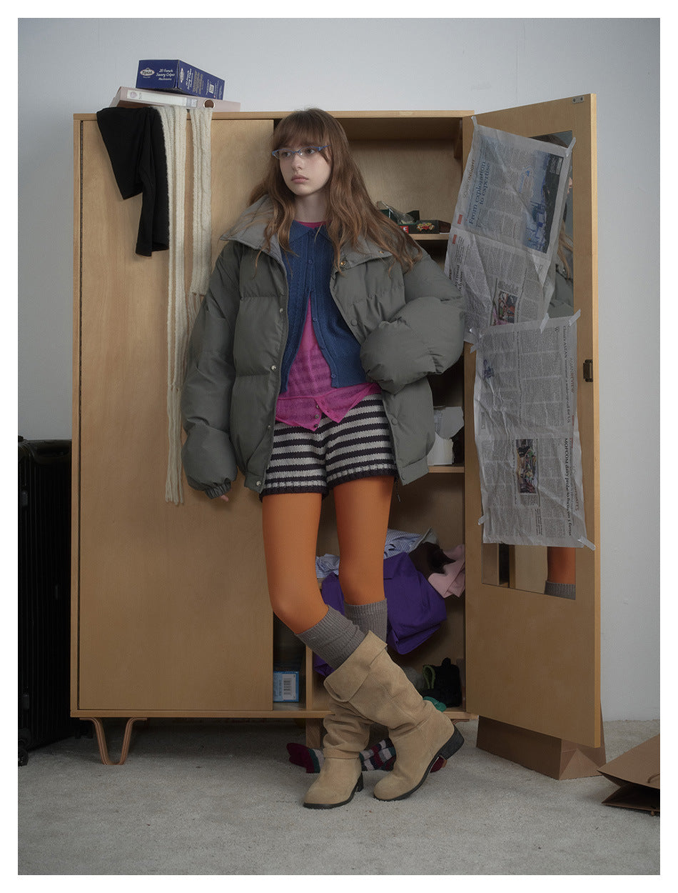Model in a gray puffer jacket with a blue cardigan, striped shorts and tan boots, posed beside an open wooden wardrobe