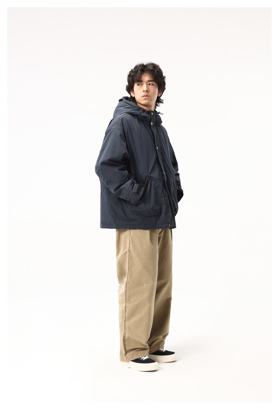 Model wearing a dark navy hooded jacket with brass-tone snaps and tan trousers, shown nearly full length.