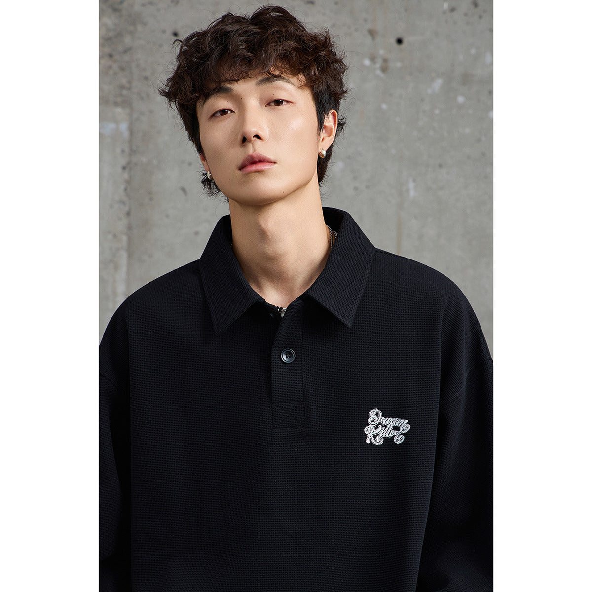 Close-up of a model wearing a black collared pullover with a button placket and small white script embroidery on the chest.