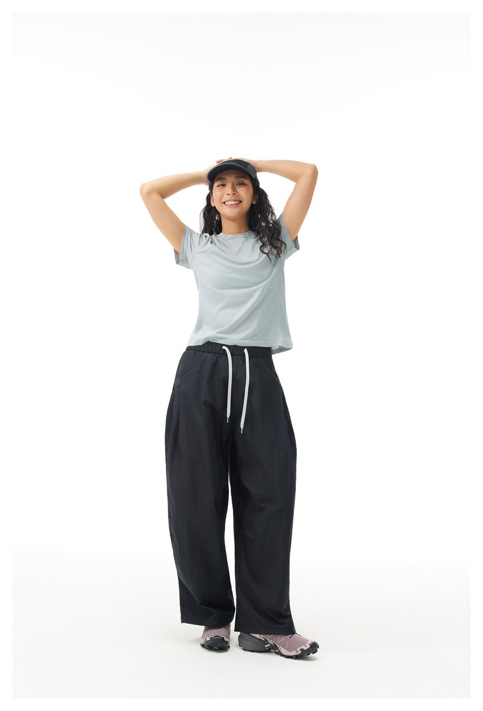Smiling model with hands on head wearing a gray-green short-sleeve T-shirt and black wide-leg pants