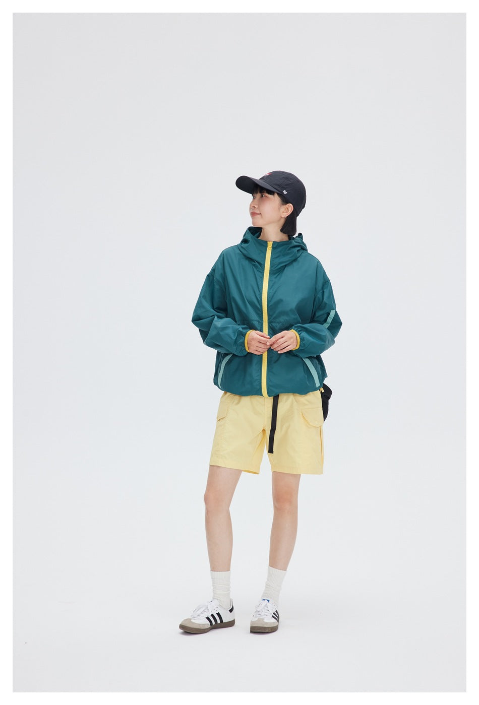 Model wearing a teal hooded zip jacket with yellow zipper and mint trim, with yellow shorts and a dark cap
