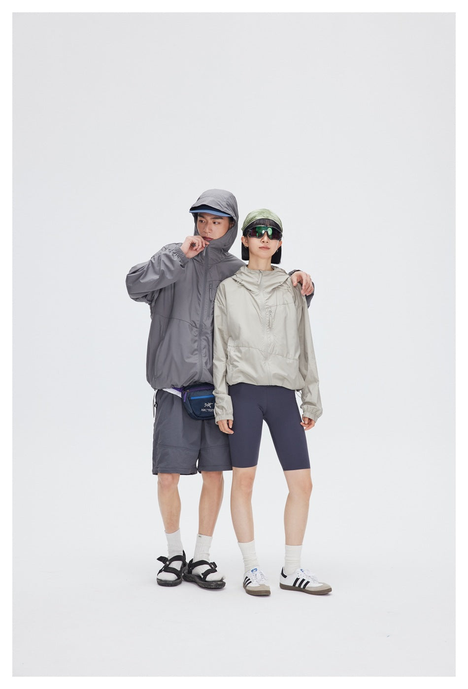 Two models wear hooded windbreakers: one gray with shorts, the other light beige with navy cycling shorts.