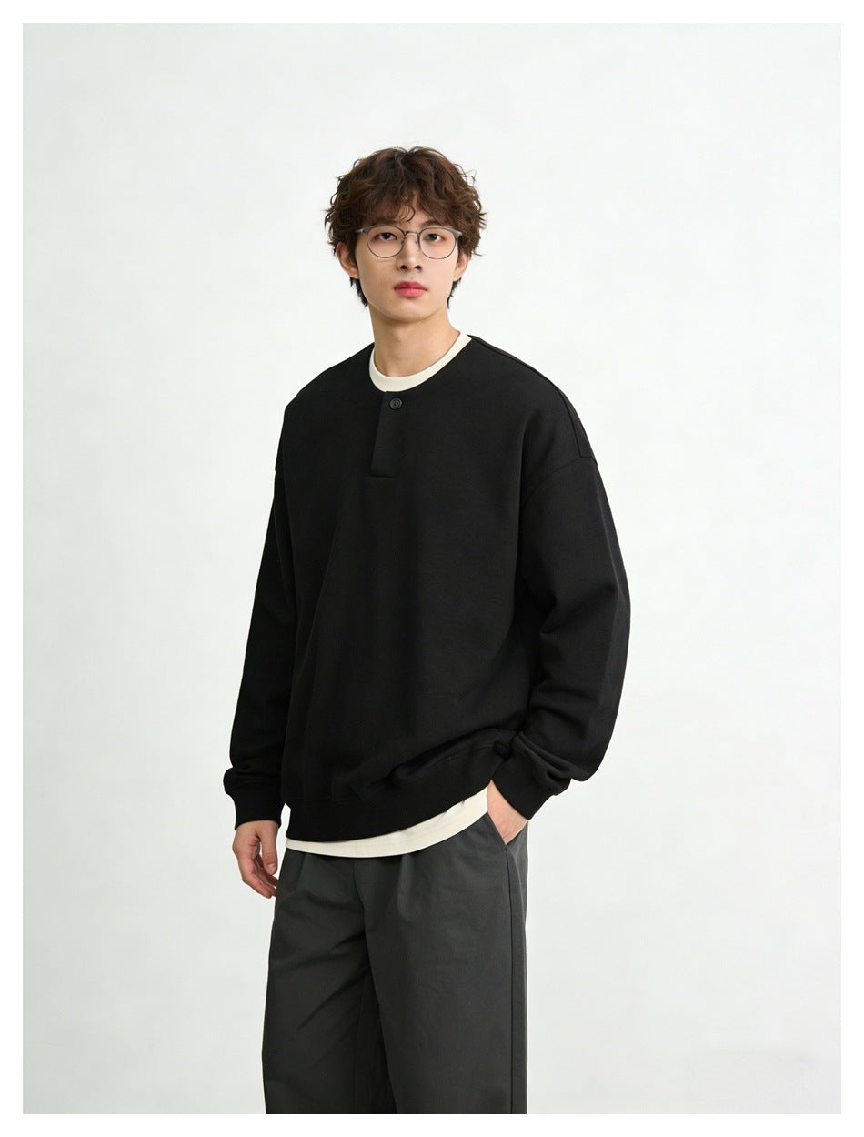 Man wearing black button-placket sweatshirt over a cream tee with gray trousers, hand in pocket, angled view