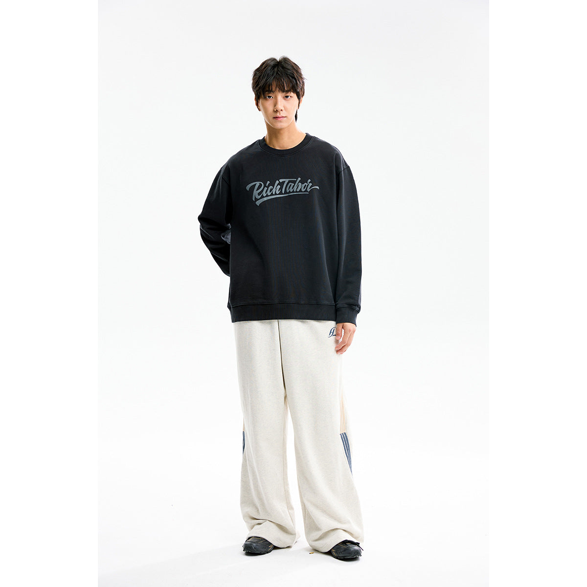Full-length model wearing a black crewneck sweatshirt with gray script print and loose light gray pants.