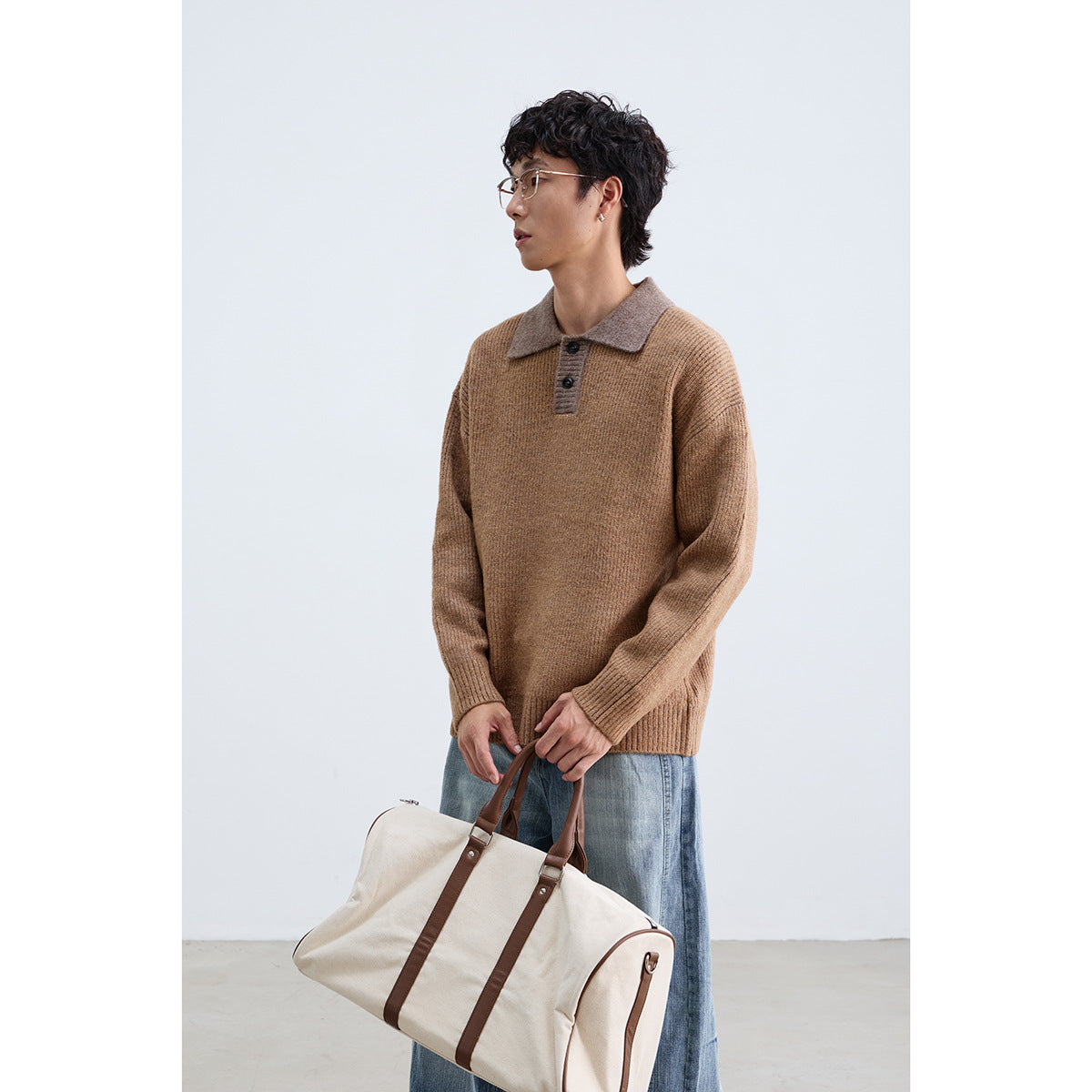 Model wearing a camel-brown knit polo sweater with a contrasting taupe collar, holding a cream duffel bag.