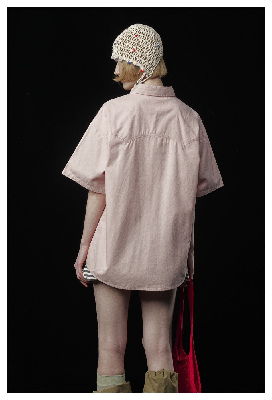 Rear view of a model in an oversized pale pink short-sleeve shirt, striped shorts, and a beaded crochet cap.