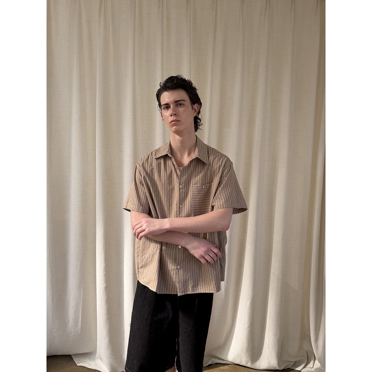 Model wearing a tan striped short-sleeve button-up shirt with a chest pocket and black trousers.
