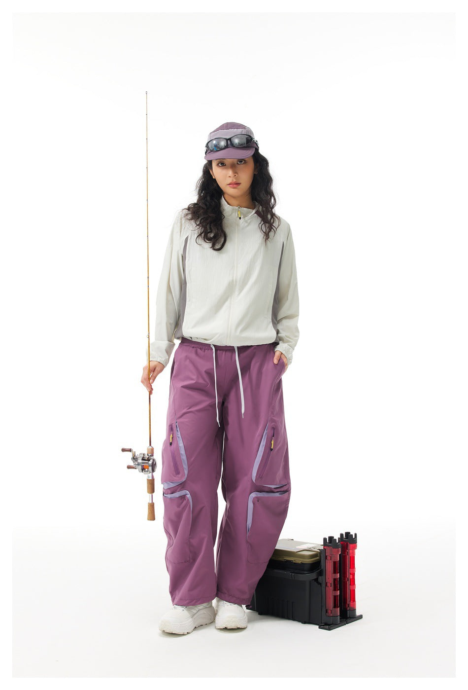Model wearing an off-white zip jacket with gray panels and plum cargo pants, holding a fishing rod.