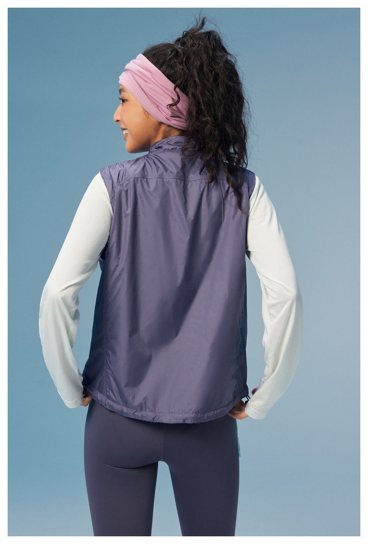Rear view of a model wearing a purple sleeveless vest over a pale long-sleeve top.