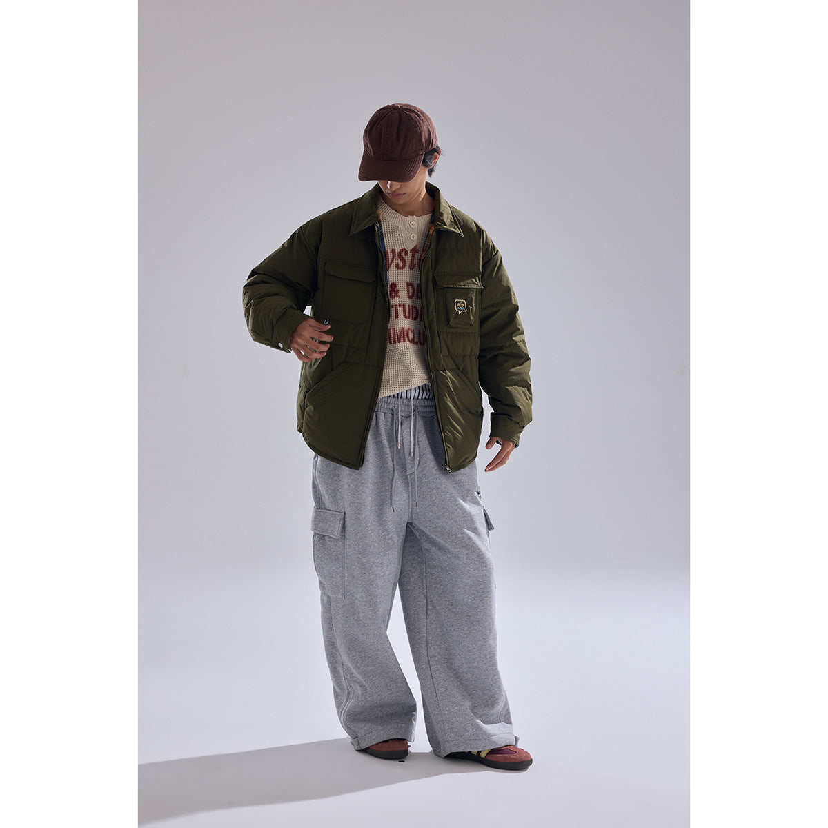 Model wearing an olive quilted jacket with flap pockets over a cream printed sweater and loose gray cargo pants.