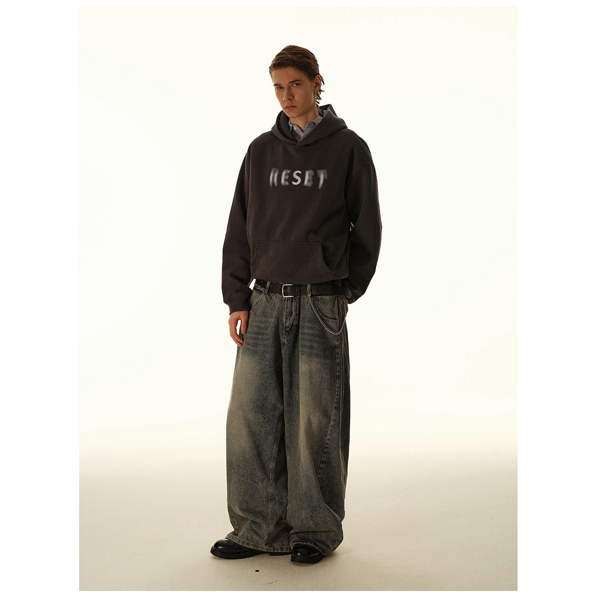 Model wearing a dark charcoal hoodie with blurred white chest lettering and oversized gray jeans.