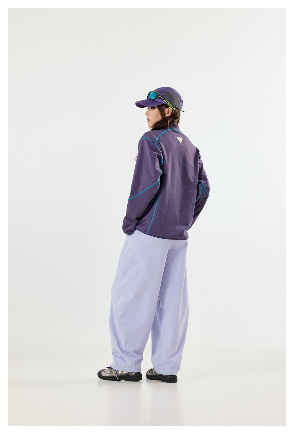 Model wearing a purple quarter-zip top with turquoise seams and loose lavender pants, shown from the back.