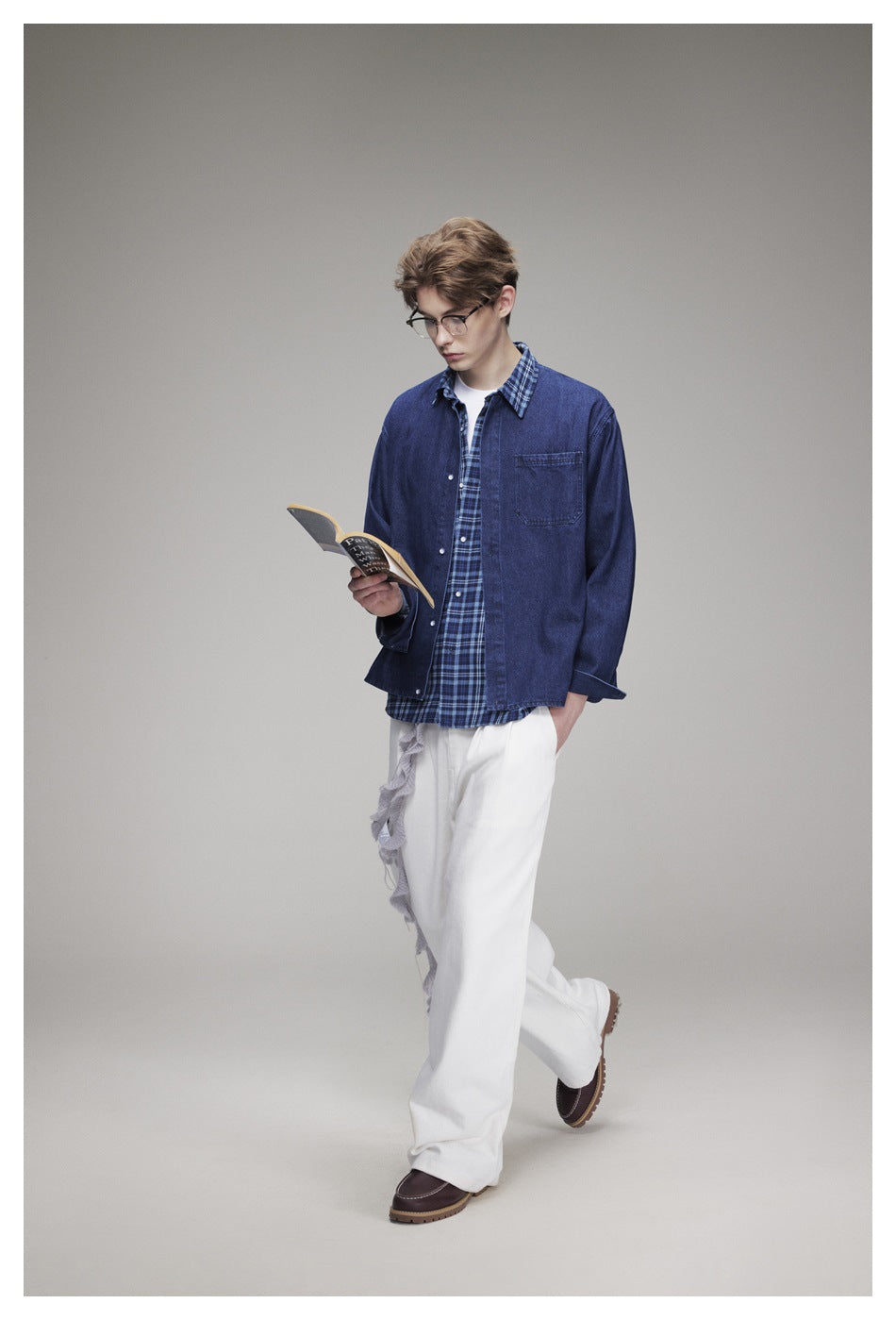 Man in blue denim shirt over plaid shirt and white pants, reading a book