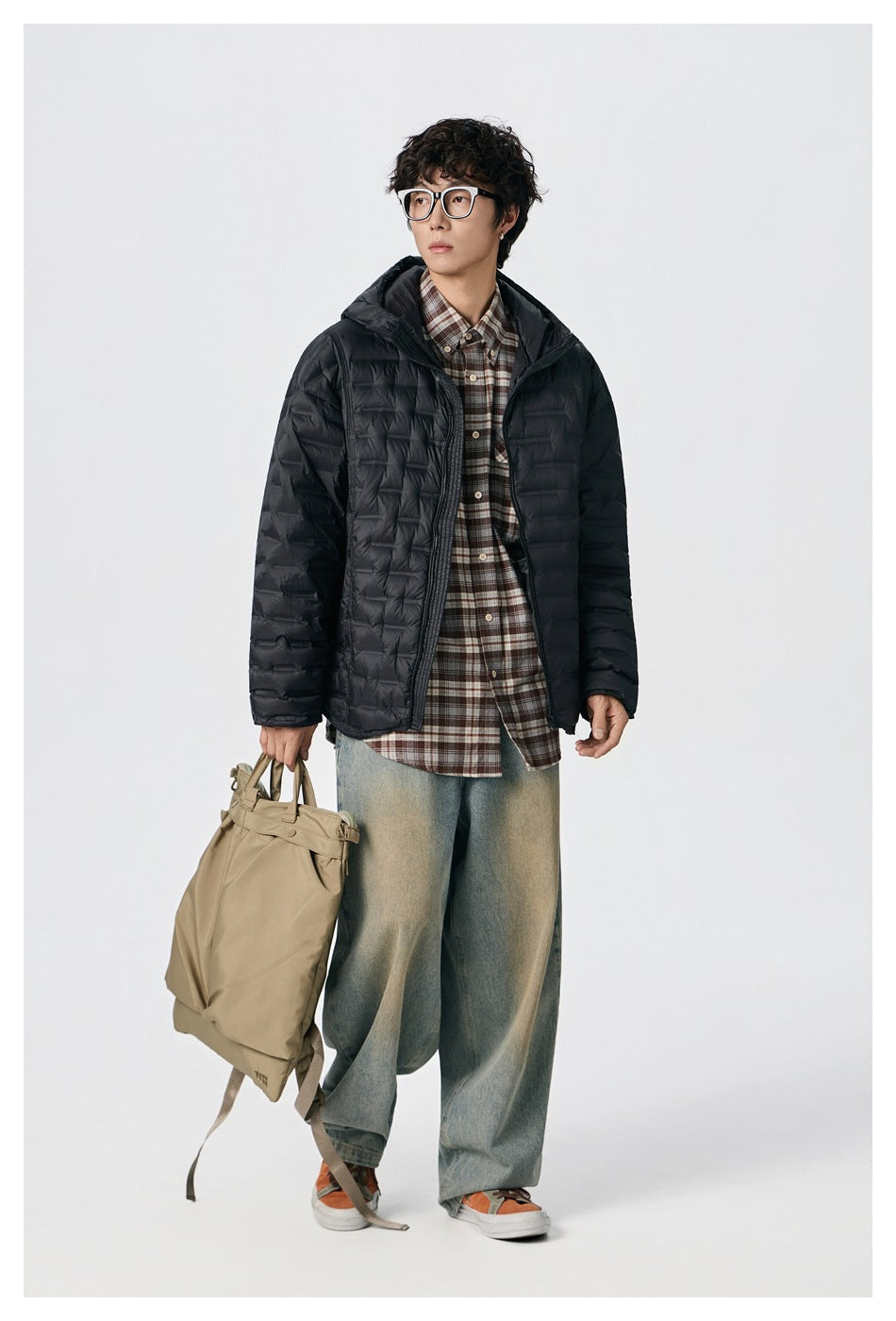 Model wearing a black quilted hooded puffer jacket over a plaid shirt, carrying a tan bag.