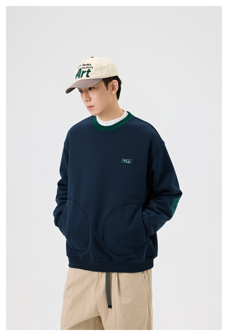 Model wearing a navy crew-neck sweatshirt with green collar, chest patch and rounded pockets, with beige trousers and a cap