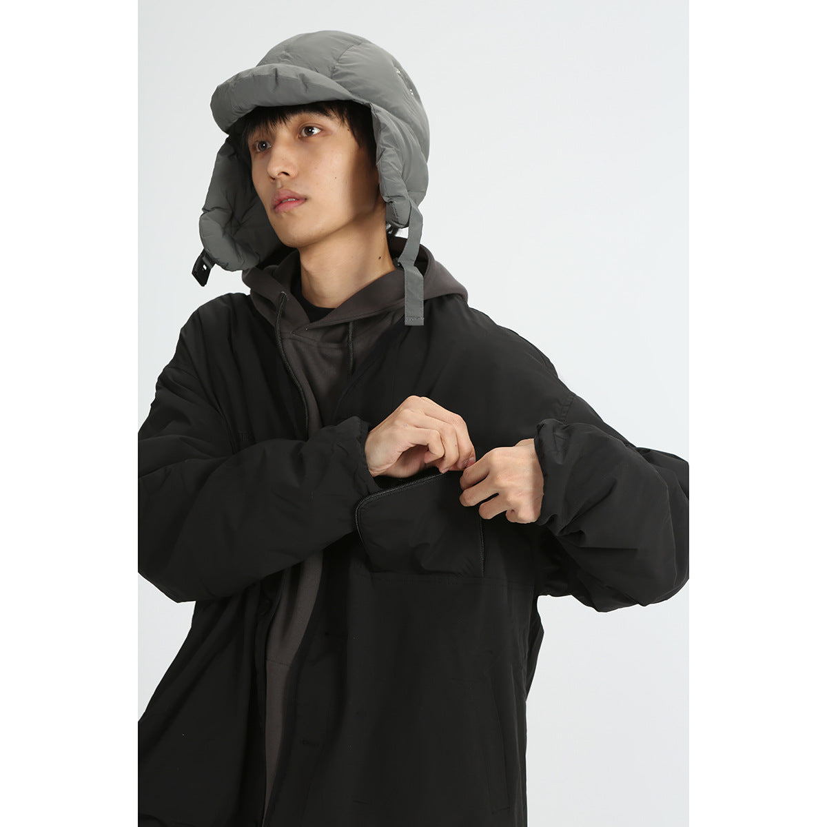 Close view of a model wearing a black jacket and gray earflap cap while opening the jacket's chest pocket.