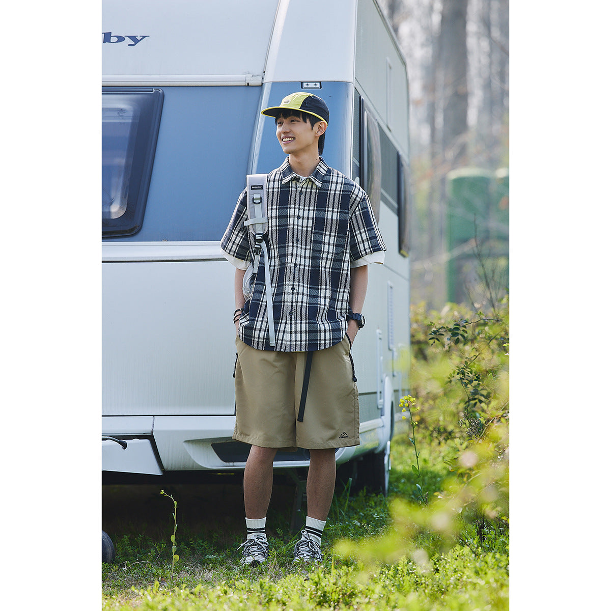 Model wearing a navy, white and gray plaid short-sleeve shirt with khaki shorts and a neon yellow cap outdoors.