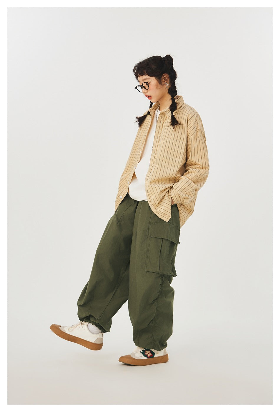 Model wearing loose olive cargo pants with flap pockets and gathered cuffs, styled with a tan striped shirt.