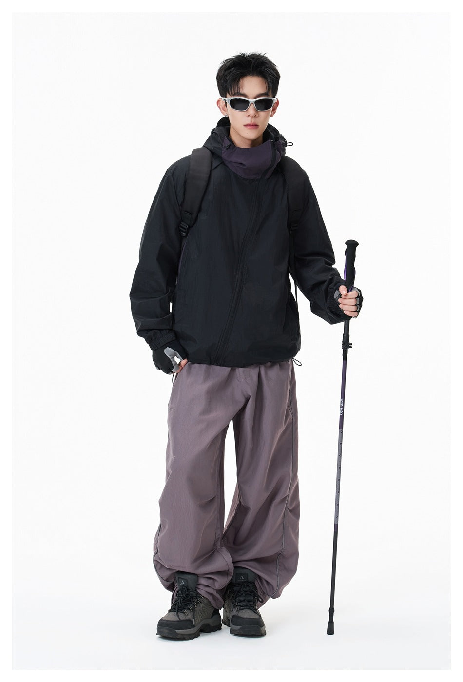 Model wearing a black hooded jacket and loose gray-purple pants, carrying a backpack and trekking pole.