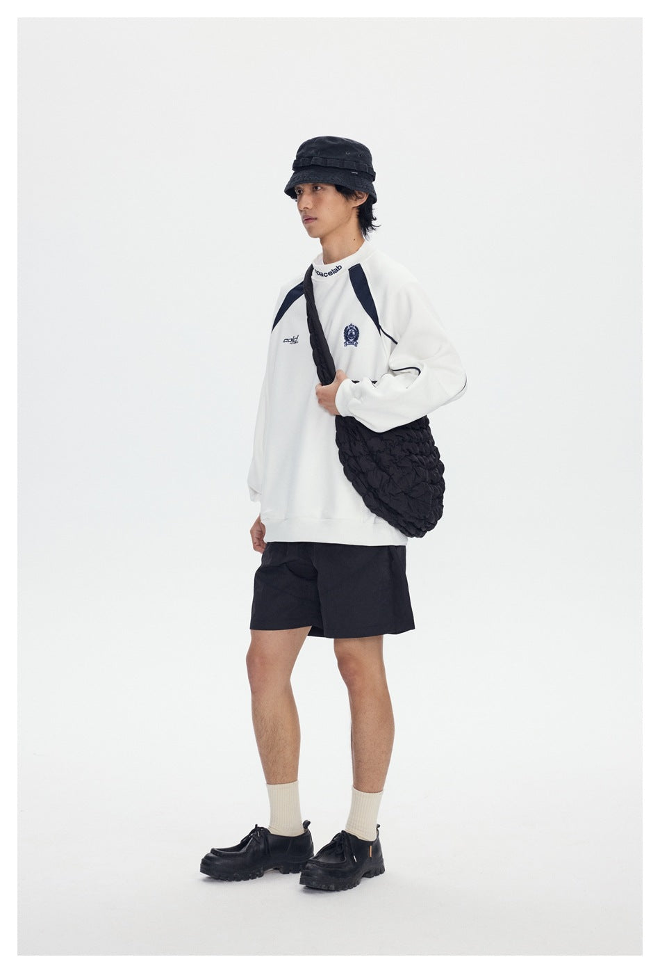 Person wearing a white crewneck sweatshirt with navy shoulder panels and piping, black shorts, bucket hat and black shoulder